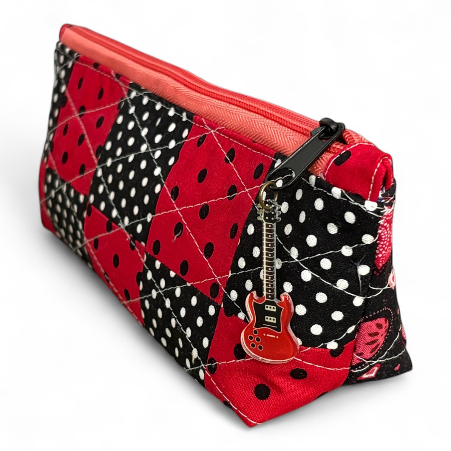 A colorful quilted pouch featuring a patchwork design with red, black, and white polka dots, topped with an orange zipper and a small guitar-shaped charm.